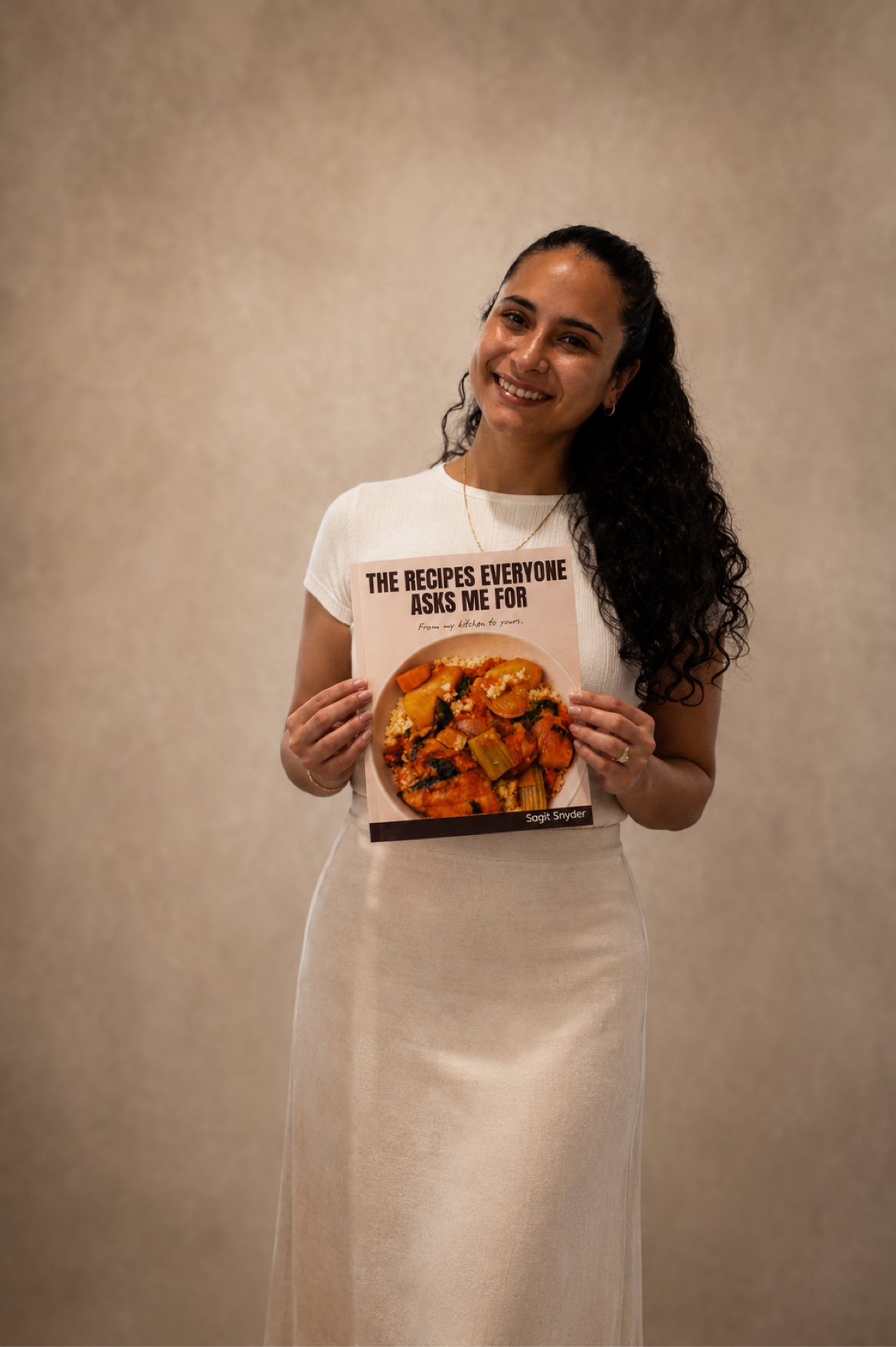 Sagit Snyder, founder of Sagit's Bakery, smiling and holding up her cookbook, The Recipes Everyone Asks Me For